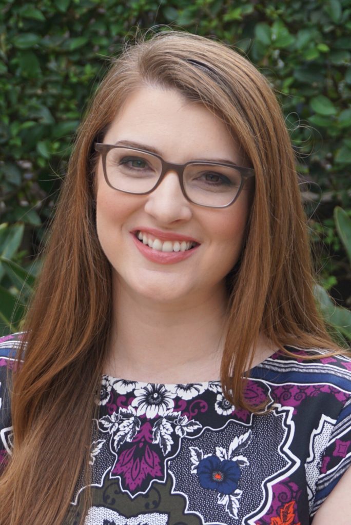 Woman with brown hair and glasses wearing colorful top in front of green background