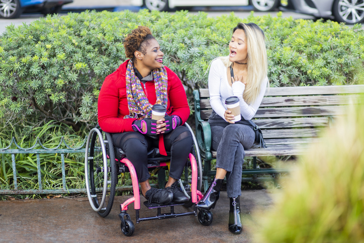 A young black disabled woman with a wheelchair and a bright colored sweater and her Asian friend walk around the city.