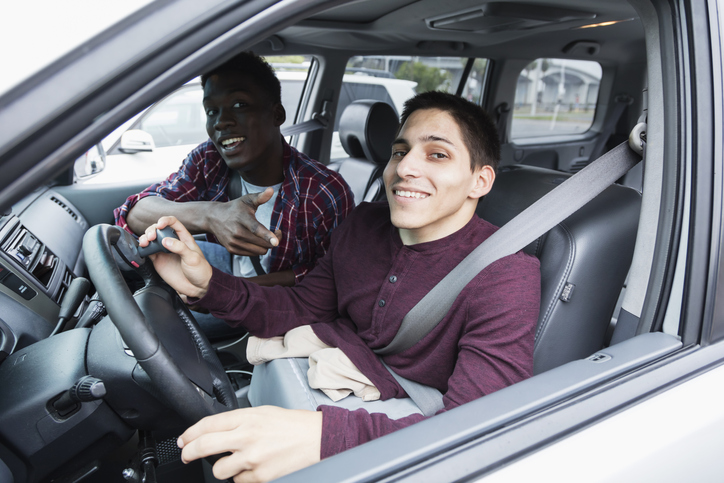 An amputee driving a car equipped with special controls on the steering wheel. He is a young man in his 20s living independently. His African-American friend is sitting in the passenger seat.