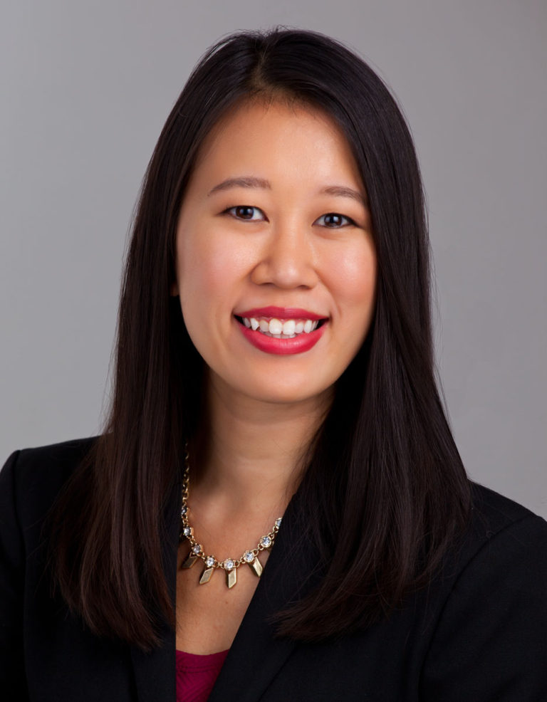 Woman with dark hair and red lipstick smiling in black blazer against grey background