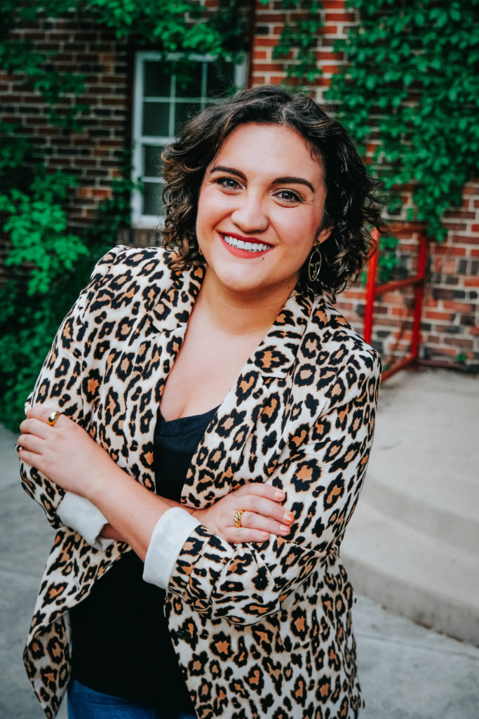 Jess, a white woman with short brown curly hair smiles at the camera. Her arms are crossed. She is wearing a leopard print blazer and black shirt. Behind her is a brick wall with green vines.