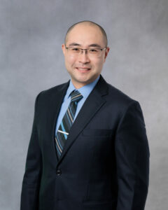 Man with short hair and glasses wearing a suitstanding in front of grey background