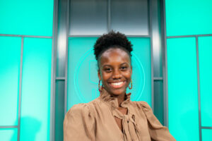 Black woman in tan suit in front of blue-green background looking at camera smiling