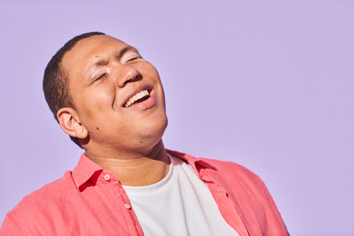 Close up portrait of happy, confident young man with autism and vitiligo standing against purple background laughing with eyes closed