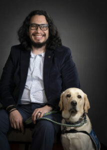 man with long hair smiling at camera wearing a white shirt and jacket sitting next to dog