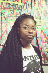 Woman with locs  and glasses looking at camera wearing black jacket and white t-shirt