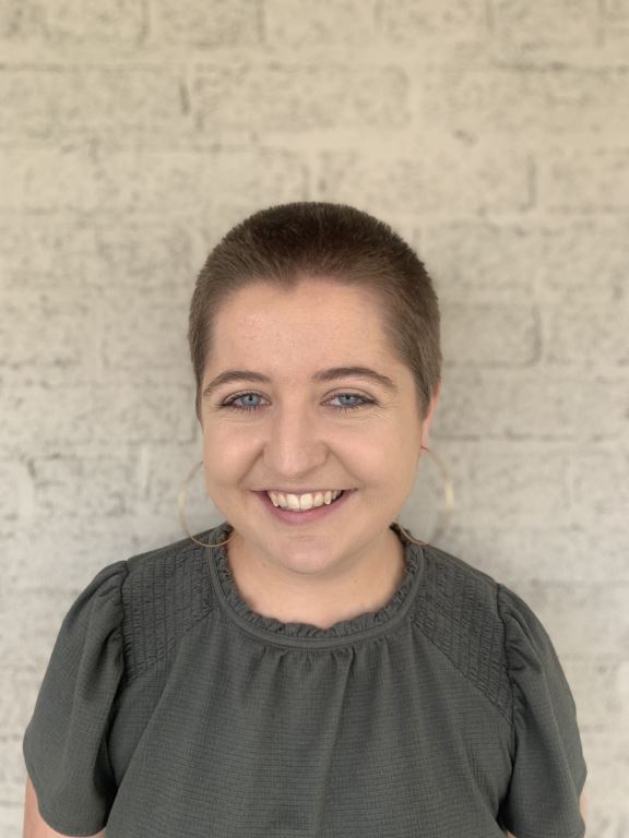 Katie is a white non-binary person with short buzz cut hair and large gold hoop earrings, smiling at the camera and wearing a dark green smocked top. They stand in front of a white brick wall.