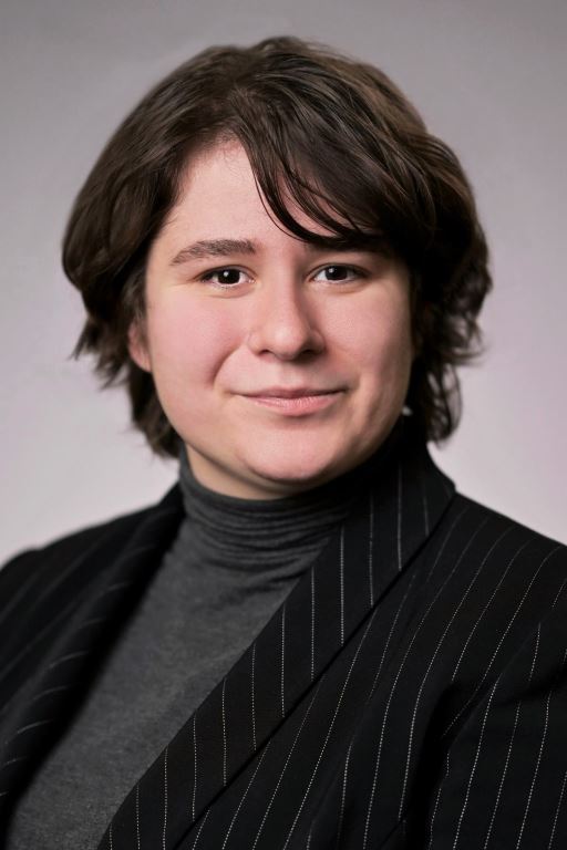 Kristen, a white nonbinary person with short wavy brown hair and sideswept bangs, smiles at the camera. They are wearing a gray turtleneck and black pinstripe blazer.