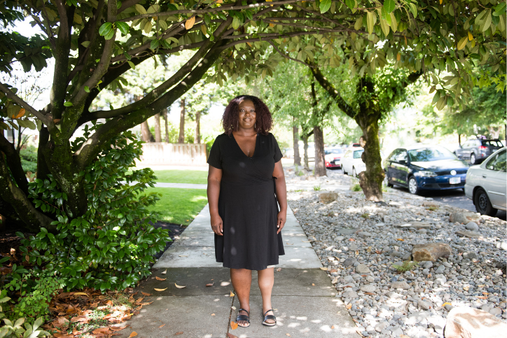 A Black woman stands under a tree and smiles, framed by green foliage all around. She is dressed in all black, with a dress and sandals and a silicone covered prosthetic leg.