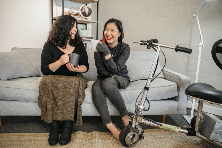 A Latinx disabled woman and an Asian disabled genderfluid person chat and sit on a couch, both holding coffee mugs. An electric lightweight mobility scooter rests on the side.