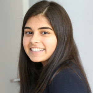 Sneha, a 21-year-old Indian woman with black is hair is smiling toward the camera inside. She is wearing a black blazer.