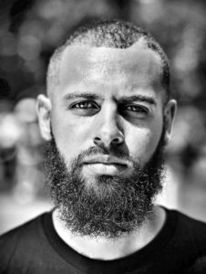 Photo of Dustin Gibson in black and white, with short hair and full beard, wearing a black round neck shirt