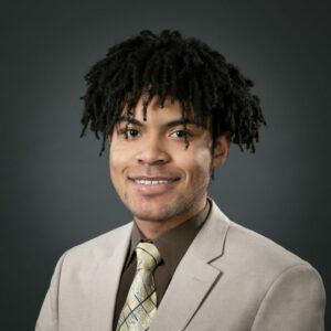 Headshot of a young Black male with small freeform dreadlocks, wearing a beige suit jacket, brown shirt, and bright yellow tie. He is smiling and facing the camera.