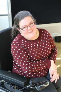 Photo of Emily Ladau smiling. She is wearing glasses and a red sweater with white polka dots.