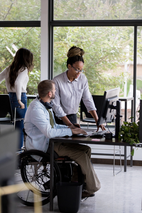 Mid adult businessman in wheelchair discusses something with a mid adult female colleague. They are using a desktop computer.