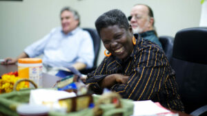 Lois Curtis, a black woman with short hair, smiles at a meeting and wears a black and yellow outfit