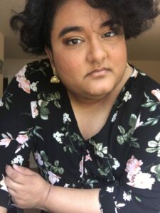 A South Asian non-binary person tilts their head. They are wearing a black floral dress and gold jhumka earrings.