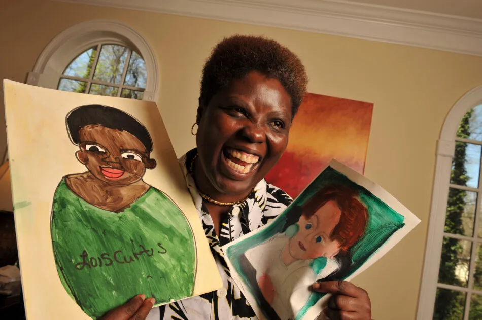 Lois Curtis, a black woman with short hair, smiles enthusiastically, and proudly holds up two portraits that she has drawn. One is a self portrait another is a portrait of a white woman with red hair