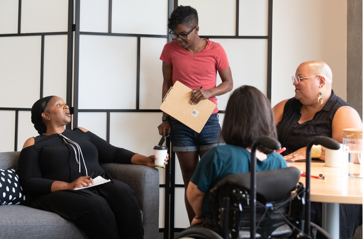 Four disabled people of color gather around a table during a meeting. A Black woman sitting on a couch gestures and speaks while the three others (a South Asian person sitting in a wheelchair, a Black non-binary person sitting in a chair, and a Black non-binary person standing with a clipboard and cane) face her and listen.