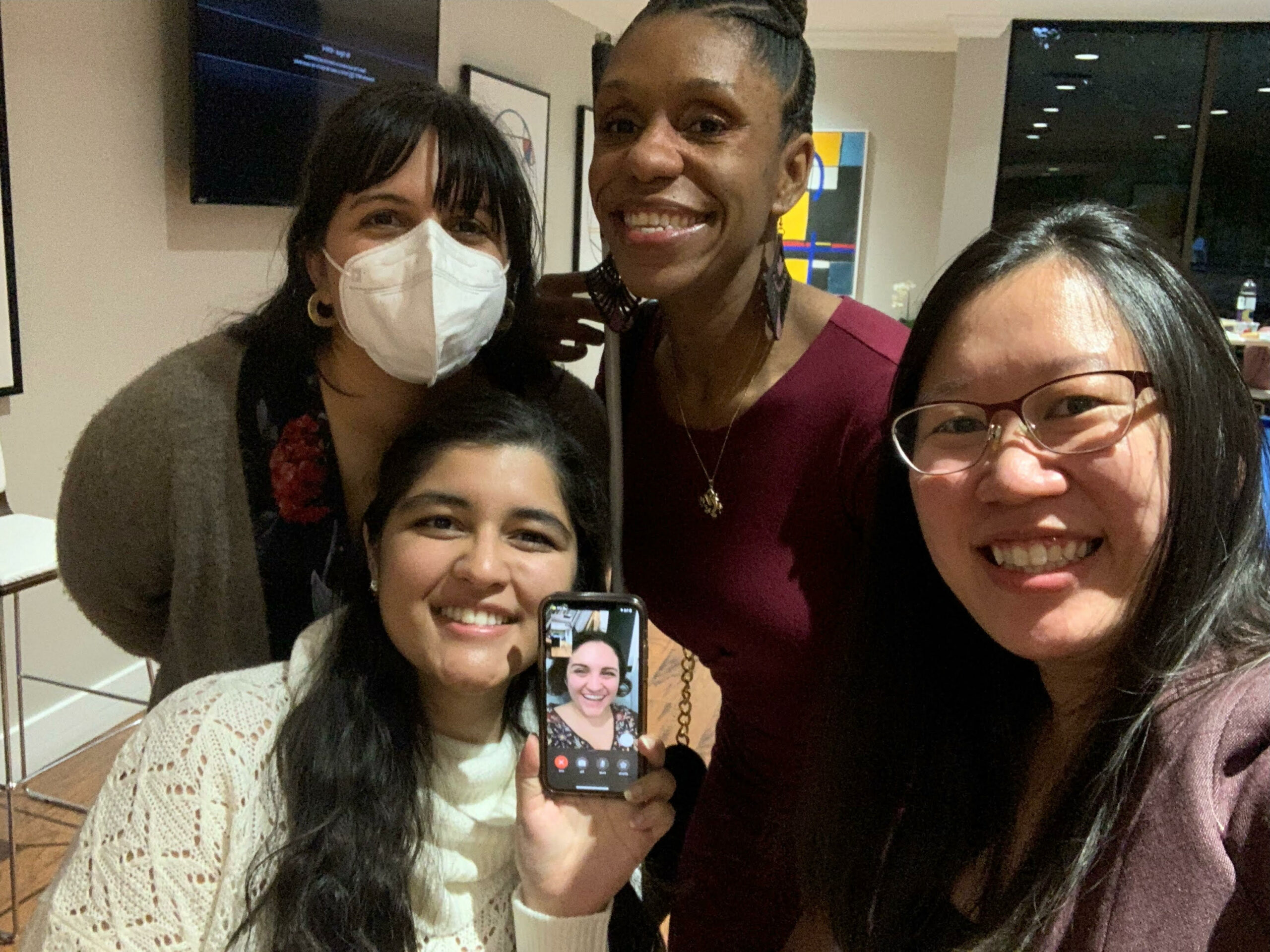 Four AAPD staff members smile at camera. One person is holding a phone with another person smiling remotely.