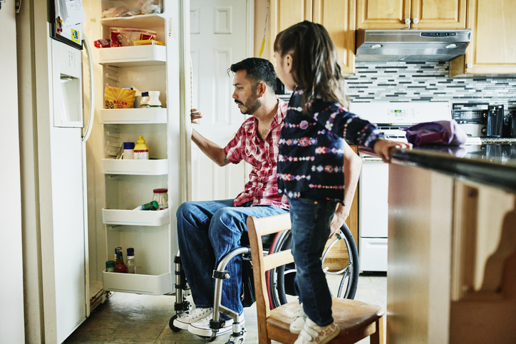 Father in wheelchair looking in refrigerator in kitchen while daughter stands on chair watching