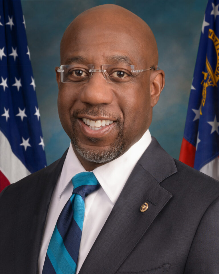 Photo of a black man smiling. He has a goatee and shaven head and is wearing rimless glasses, a blue and teal striped tie, white dress shirt, and charcoal gray suit. In the background is the American flag and the Georgia state flag.