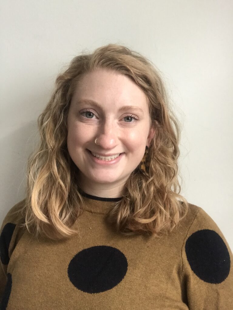 alt="A young white woman with curly blond shoulder-length hair smiling at the camera while wearing brown sweater with black polka dots and a nose ring. She is standing in front of a plain white wall."