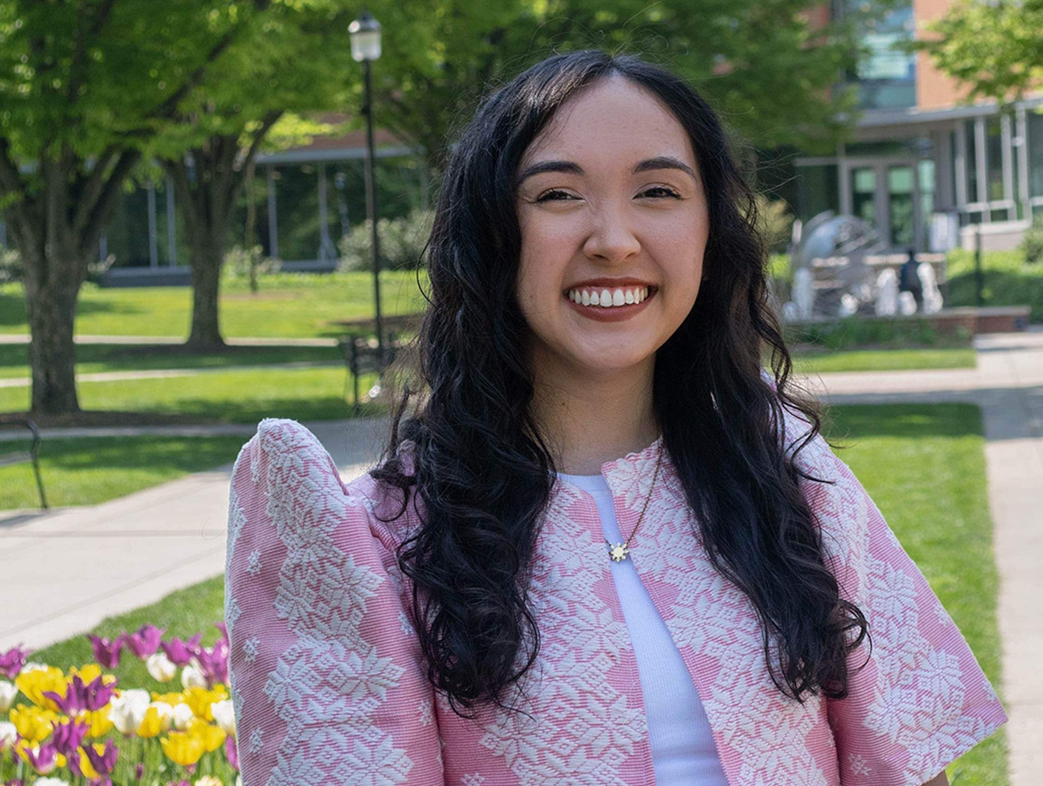 April is standing outside in Vartan Plaza at Penn State Harrisburg. She has curly, dark brown hair and is wearing a pink and white woven bolero, a white shirt, and a gold Philippine sun necklace. Yellow, white and purple tulips are in full bloom.