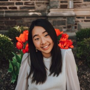 Headshot of Jennifer Lee. Jennifer is an Asian woman with long black hair wearing a cream blouse with sheer sleeves. Jennifer is in front of a brick building with bright orange flowers behind her.