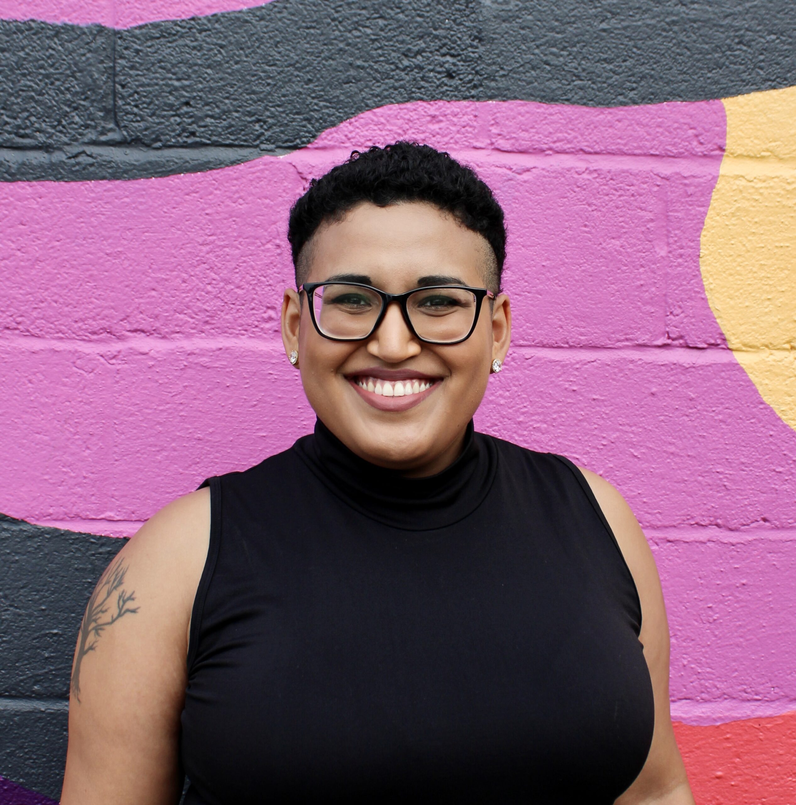 Afro-Latinx person of medium brown complexion with short curly black hair smiling. They are wearing a black turtleneck tank top and black eyeglasses and pink lipstick, against a vibrant and colorful background of pink and green hues.