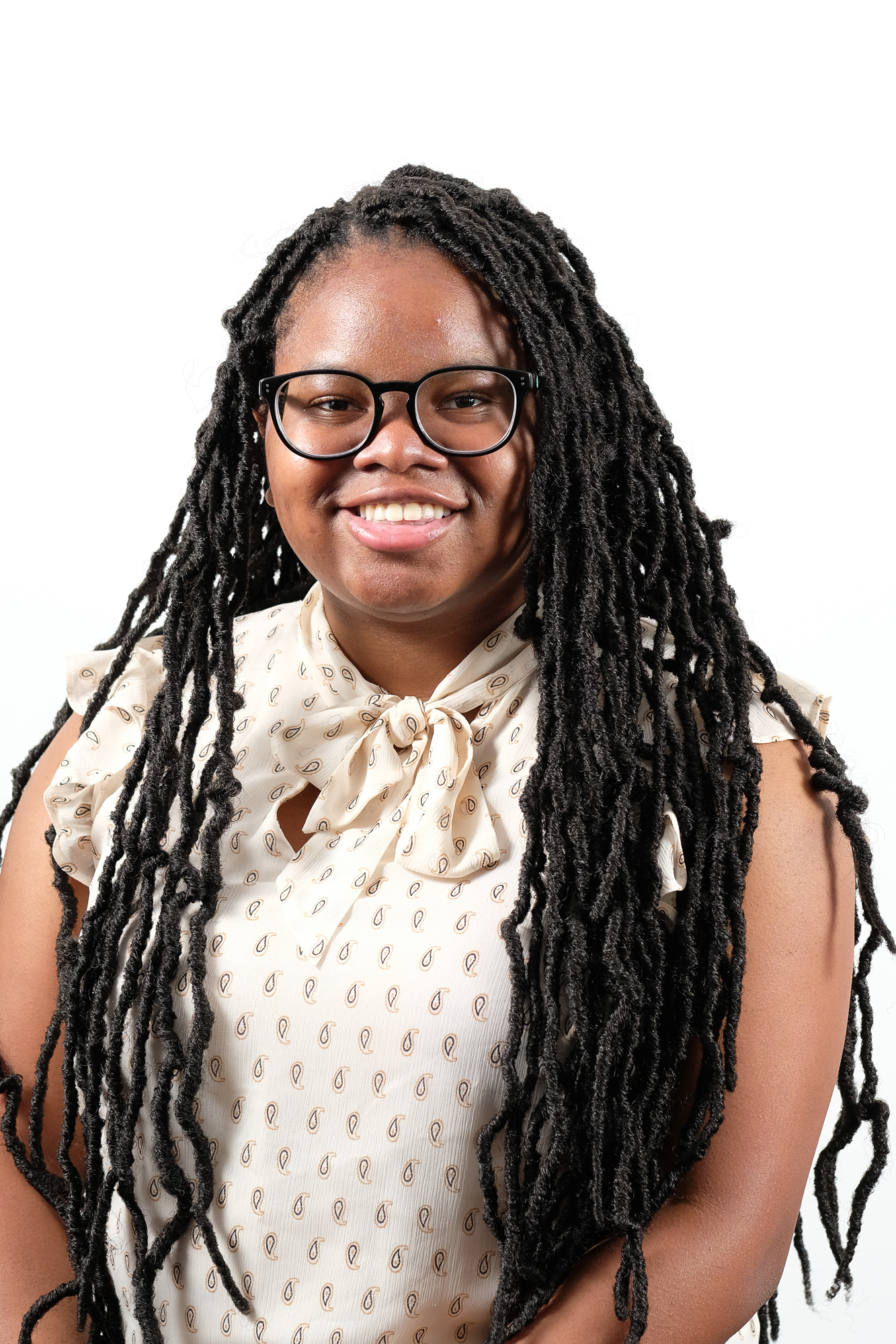 Headshot of Christina Stafford, a black girl, from shoulders up, with her hair in locs. She is wearing glasses and a white patterned top.