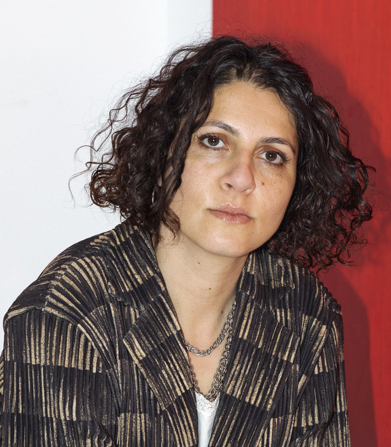 Headshot of Josephine Shokrian, a light skin brown person with chin length brown curly hair posing in front of a red and white background wearing a black and brown blazer with white camisole.
