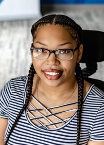 Headshot of Keisheona, a woman with  light brown skin, hair braided back in two braids, wearing black framed glasses, and a navy blue and white horizontal stripe shirt.