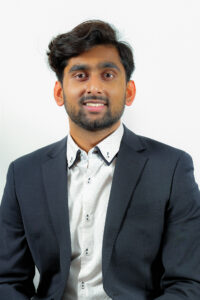 A headshot of Kinshuk smiling at the camera. He is wearing a charcoal blue suit with a dotted white dress shirt. Kinshuk has medium length hair in a side swept quiff hairstyle and a stubble length beard.