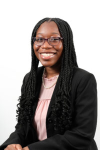 Headshot of Lauren Proby, a young African American woman with black knotless braids, smiling warmly at the camera. She is wearing dark purple glasses and a pink blouse covered with a black suit jacket. She also wears a string of pearls.