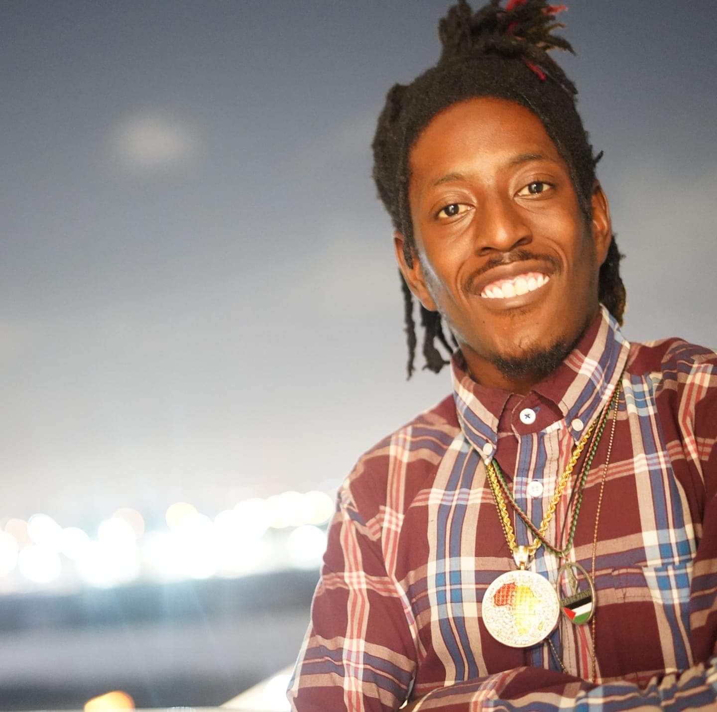 Standing just right of center stands a Black man smiling with teeth showing. The background is of a city lit up at night. His arms are folded and he is wearing a maroon colored shirt with a plaid pattern that has blue streaks in it. He has two chains, one gold of the continent of Africa another of the flag of Palestine . He has his hair styled in loc form and is wearing them up in the shape of a pineapple. There are green and red tips in his hair.