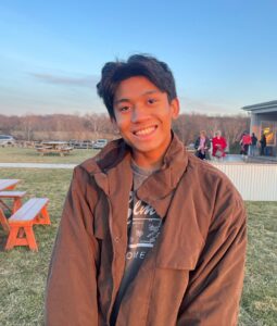 Headshot of Logan Jalil, tan-skinned Asian male, with short black hair, wearing green jacket and grey t-shirt. The background is outside during a sunset.