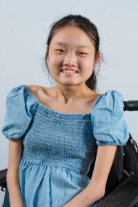 Headshot of Melissa Shang, a Chinese-American woman with black hair tied in a ponytail. She is wearing a denim blue dress and sitting in a power wheelchair. She is smiling. In her background is a white wall