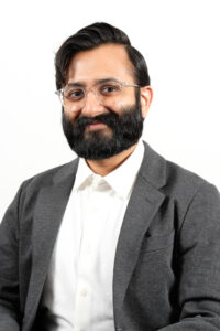 Headshot of Neil Purohit, a South Asian man with short, side-parted hair, a beard, and glasses, wearing a white button down shirt and grey suit jacket, in front of a bright, plain white background.