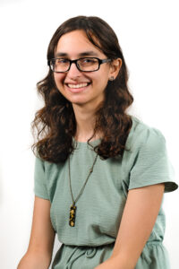 Headshot of Rachel Litchman, a white person with long, wavy brown hair. She is wearing black glasses, a light green shirt, and a necklace with a yellow and black crystal.