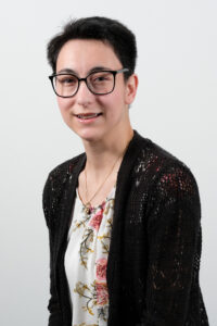 Headshot of Sarah Smith, a Latina female with short black hair, black rimmed glasses, and  braces. She is wearing a white floral top and black cardigan.