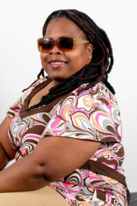 Headshot of Shariese Katrell, a black female with locs, wearing sunglasses, and a retro brown, pink, orange, grey, and white shirt.