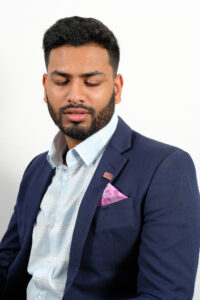 Headshot of Shawn Abraham, an Indian man with a trimmed beard, wearing a blue and white checkered button up and a navy suit jacket with a pink pocket square.