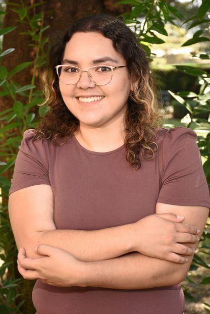 Shirley is a Hispanic woman with brown hair, wearing a purple blouse and clear glasses. She is smiling at the camera with her arms crossed over her chest standing in front of a tree