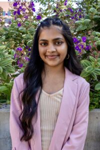 Headshot of Shreya Singh, a brown-skinned South Asian woman with long black hair. She wears a pink blazer and cream colored shirt. 