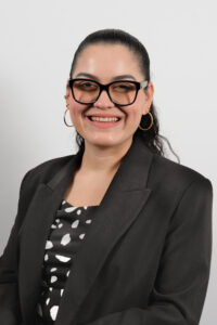 Headshot of Stephanie Picazo, a light skinned Latina with black curly hair pulled back into a ponytail. She wears black glasses, gold hoop earrings, a black and white patterned top and black blazer.