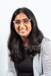Headshot of Trisha Kulkarni, with her hair down. She is wearing stylish glasses and a black dress and grey cardigan.