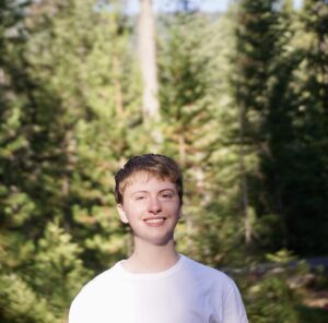 Elian, a white man with short, light-brown hair, is wearing a white t-shirt and is smiling outside on a bright, sunny morning. Behind him are a number of towering Douglas fir trees. 