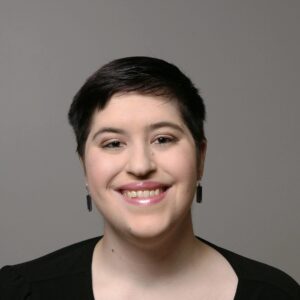 Kaeli, a white woman with short brown hair against a gray backdrop. She wears dangling, purple, earrings and smiles at the camera. She also wears a black shirt.