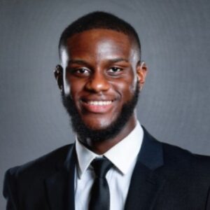 This headshot depicts Oluwaseyi Onifade, a young black man wearing a black suit, white shirt, and black tie. He is smiling in the photo.